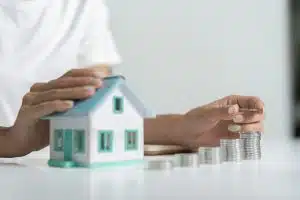 Person Counting Coins Next to a Small Model House on a White Table, Concept of Saving Money for Real