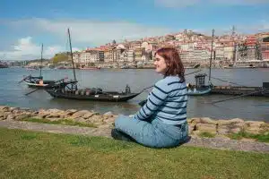 Young woman sitting on riverside in Porto, Portugal overlooking traditional boats and cityscape